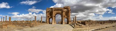 Tall single-bay Roman arch centered on a paved ancient route, with columns and low ruins extending on both sides at Timgad.