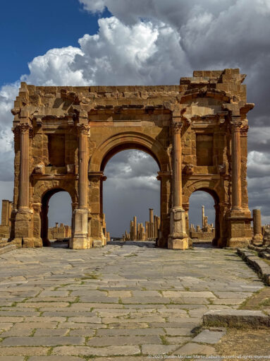 Broad frontal view of Timgad’s Arch of Trajan with one large central arch, two smaller side openings, columns, and ancient paving.