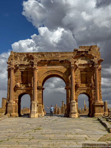 Visitor standing beneath the Arch of Trajan at Timgad, framed by the large central opening, two side arches, and paved street.
