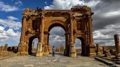Close oblique view of Timgad’s Arch of Trajan with central vault, side passage, engaged columns, and a visitor beneath the arch.