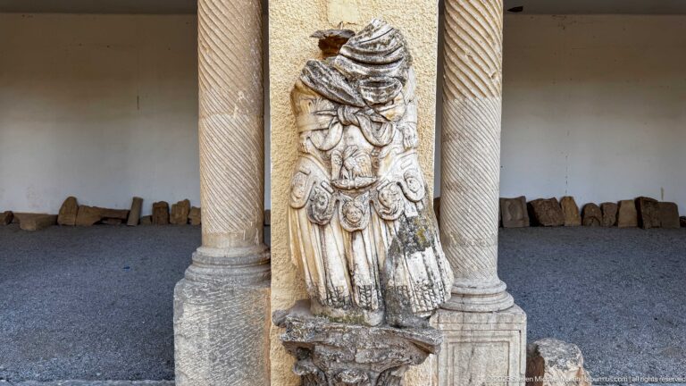 Weathered carved stone figure displayed between two spiral-fluted columns, with additional stone fragments arranged along the wall.