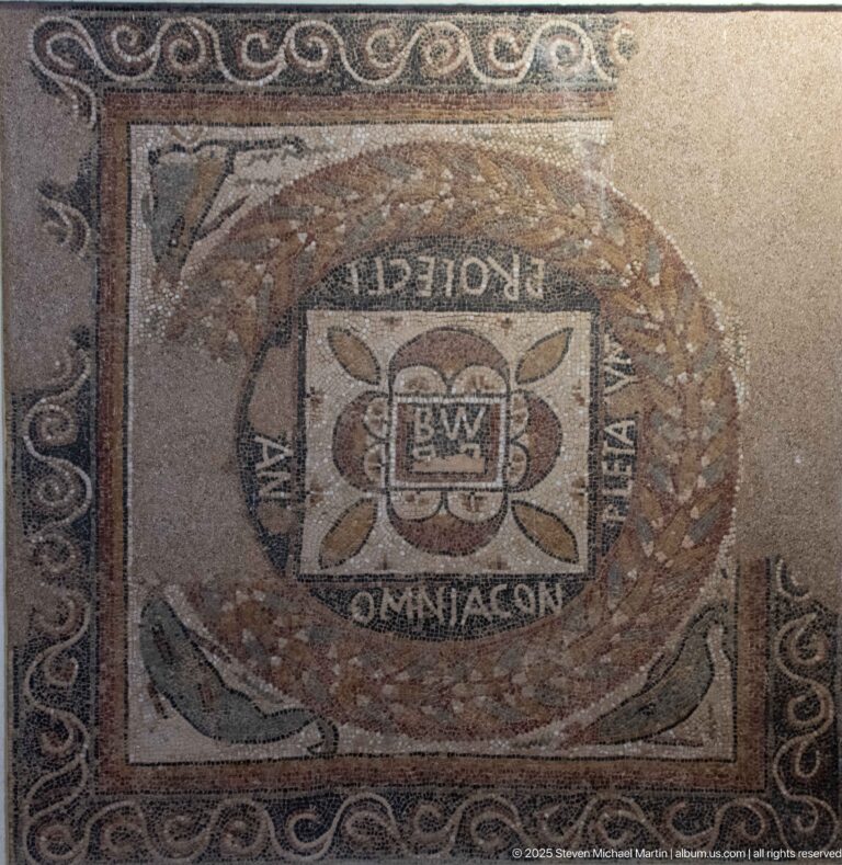 Inscribed Roman mosaic with a square central emblem, oval wreath, scrolling border, and partially legible Latin text.