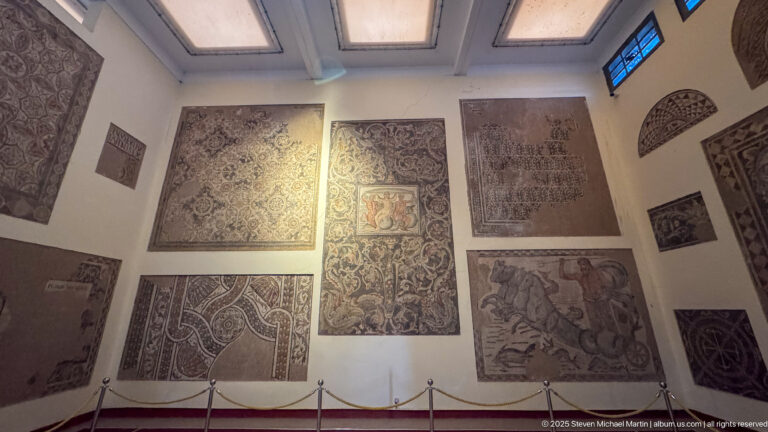 Museum gallery wall displaying numerous mounted Roman mosaic panels of different sizes, patterns, and subjects.