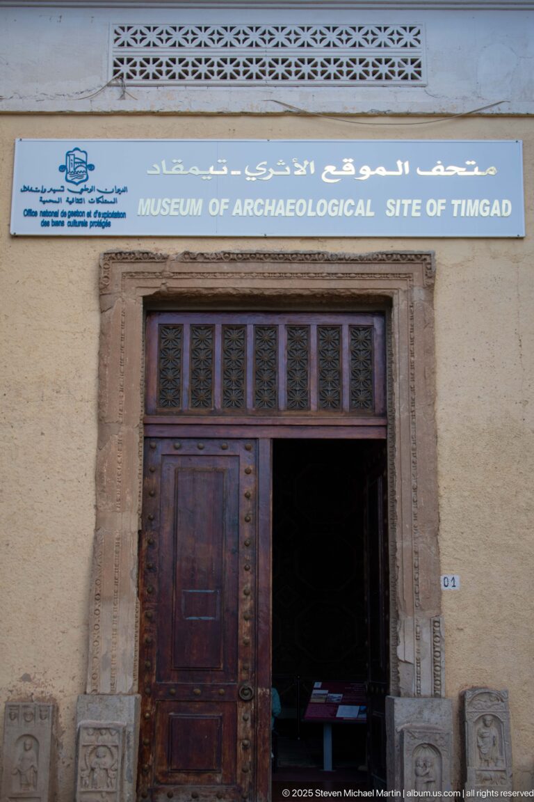 Entrance to the Archaeological Museum of Timgad with bilingual sign, wooden doors, and carved stone fragments beside the doorway.