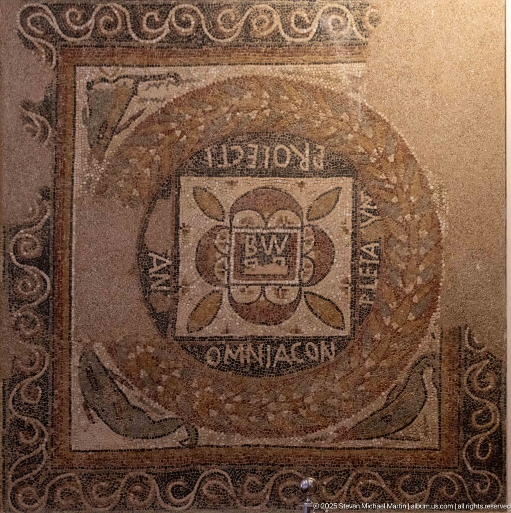 Roman inscribed mosaic with square central emblem, oval wreath, surrounding Latin lettering, and scrolling borders.