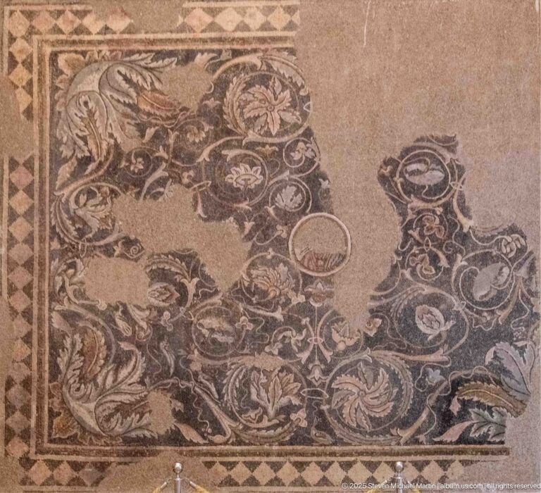 Fragmentary Roman mosaic with scrolling foliage, birds, floral motifs, circular vessel-like forms, and a large central loss.