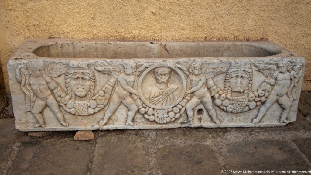 Carved rectangular stone funerary object with garlands, masks, and a central circular portrait medallion.
