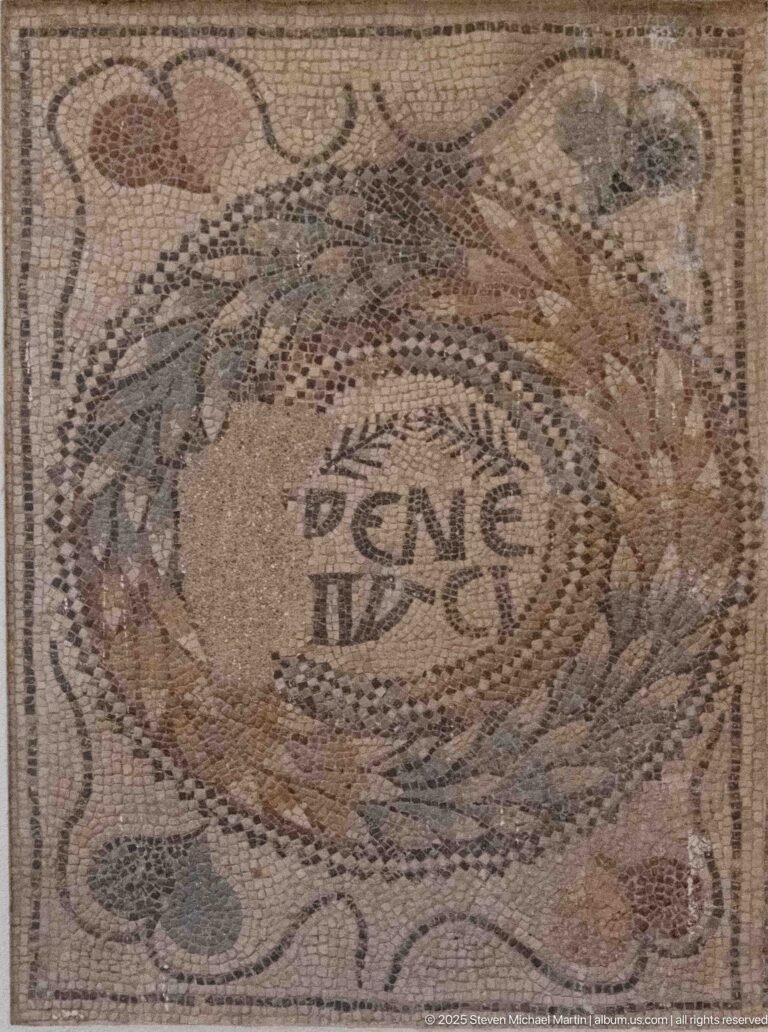Roman mosaic medallion with a short Latin inscription surrounded by curving colored ornament.