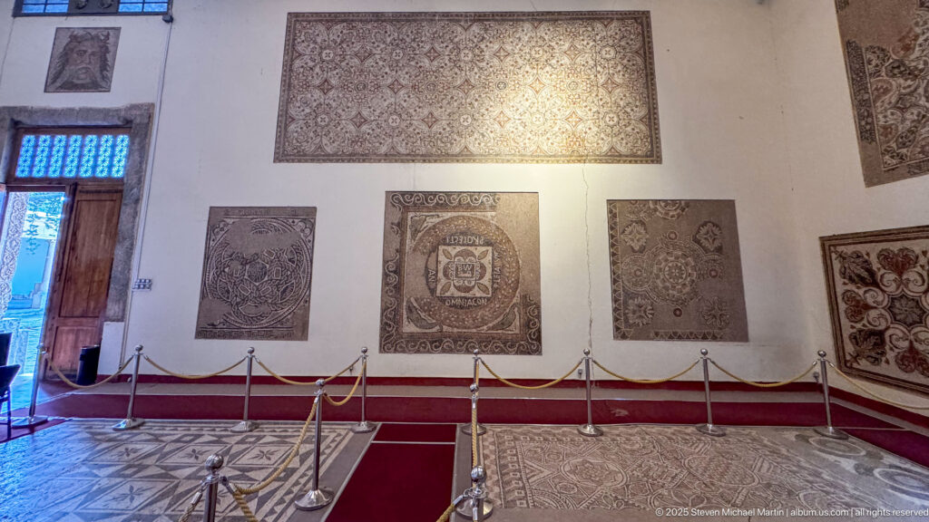 Museum gallery with several Roman mosaics mounted on cream walls and a large floor mosaic protected by ropes.