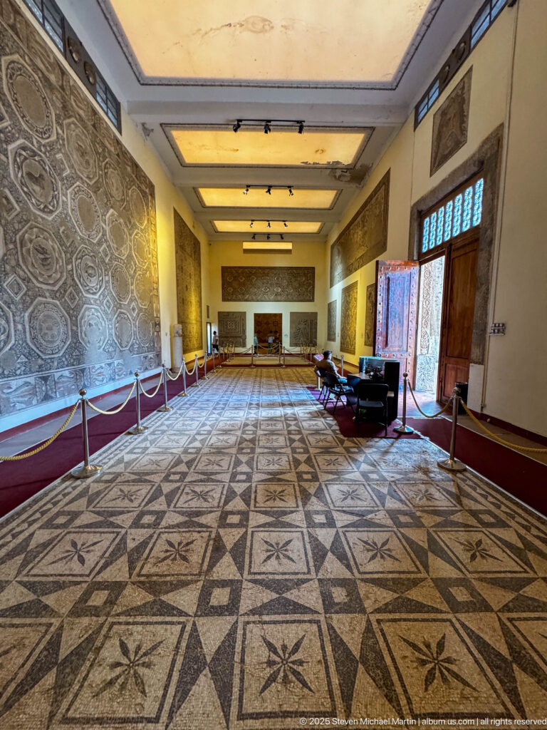 Long museum gallery with a geometric mosaic floor, wall-mounted mosaics, high windows, and display barriers along both sides.