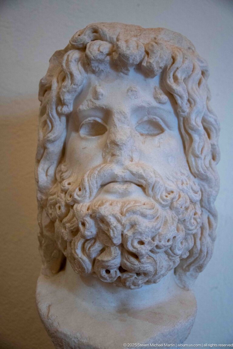 Close view of a bearded marble male head with deeply carved curls, heavy brows, and a weathered broken neck.