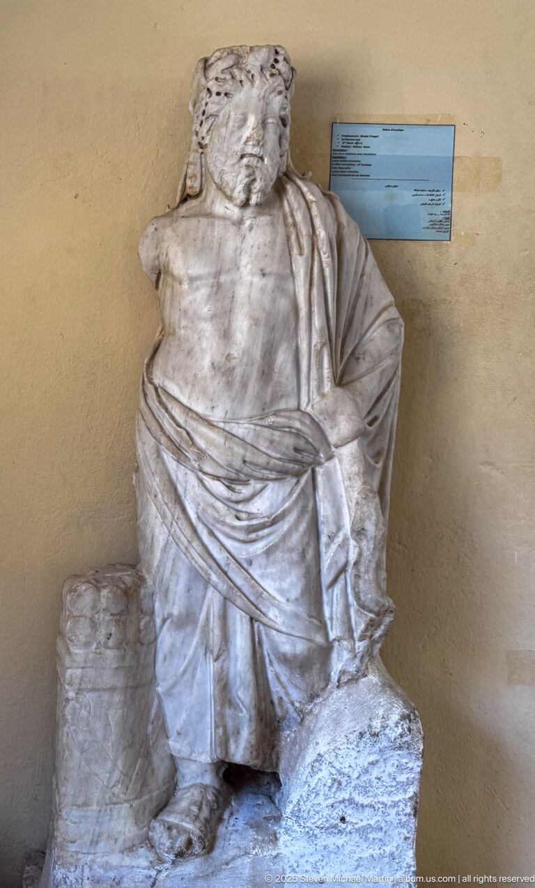 Fragmentary seated marble male statue in heavy drapery, displayed against a wall with a museum label behind it.