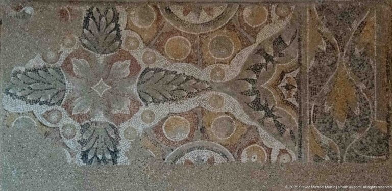 Fragmentary polychrome mosaic with a large pale flower, circular motifs, curved borders, and areas of loss.