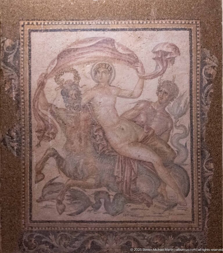 Square Roman figural mosaic with a standing central figure, smaller flanking figures, and an ornate decorative border.