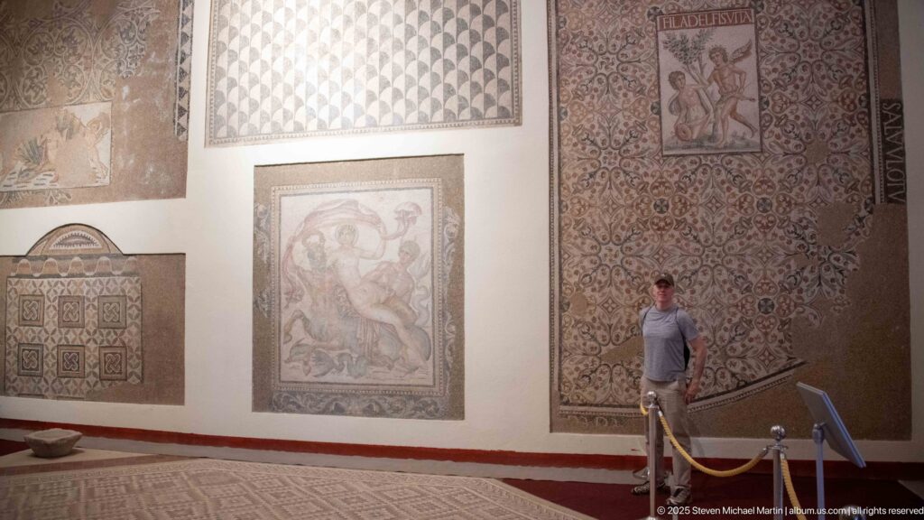 Visitor standing beside several large wall-mounted Roman mosaics, including the tall panel inscribed FILADELFIS VITA.