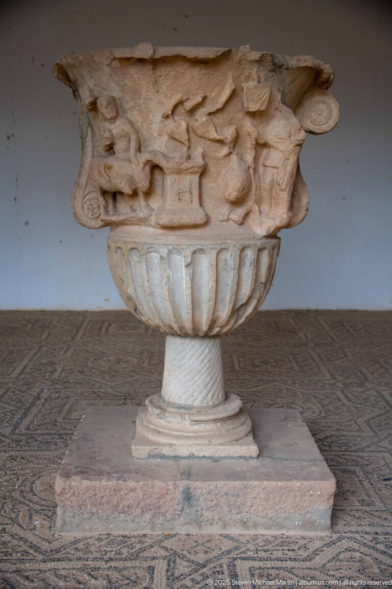 Close view of a marble basin or urn with small carved human figures around the upper relief and deep fluting below.