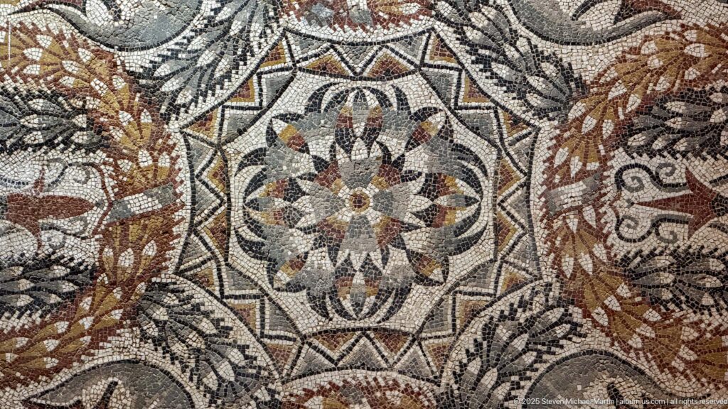 Close-up of a polychrome mosaic with a multi-petaled central rosette, angular frame, and surrounding foliage.