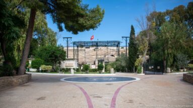 Paved landscaped approach to the Roman Theatre of Carthage with stone seating, stage structure, trees, and Tunisian flag.