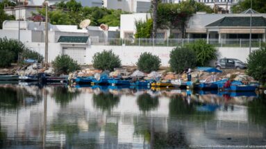 Row of small fishing boats, nets, and gear along the Punic port shoreline below white residential buildings.
