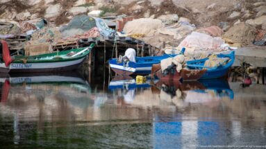 Blue and green fishing boats moored beside piled nets, tarps, and gear, with a person working at the shoreline.