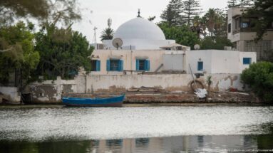 White waterfront house with blue shutters, large white dome behind, and a small blue boat moored on the Punic port basin.