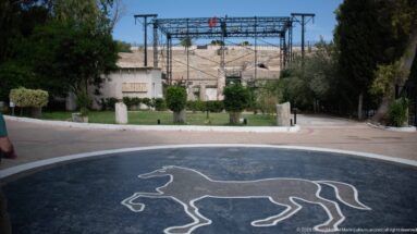 White outline of a horse on dark circular pavement with the Roman Theatre of Carthage beyond.