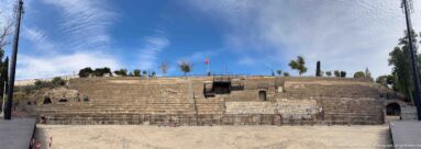 Wide frontal view of the Roman Theatre of Carthage with curved stone seating, access openings, and Tunisian flag.