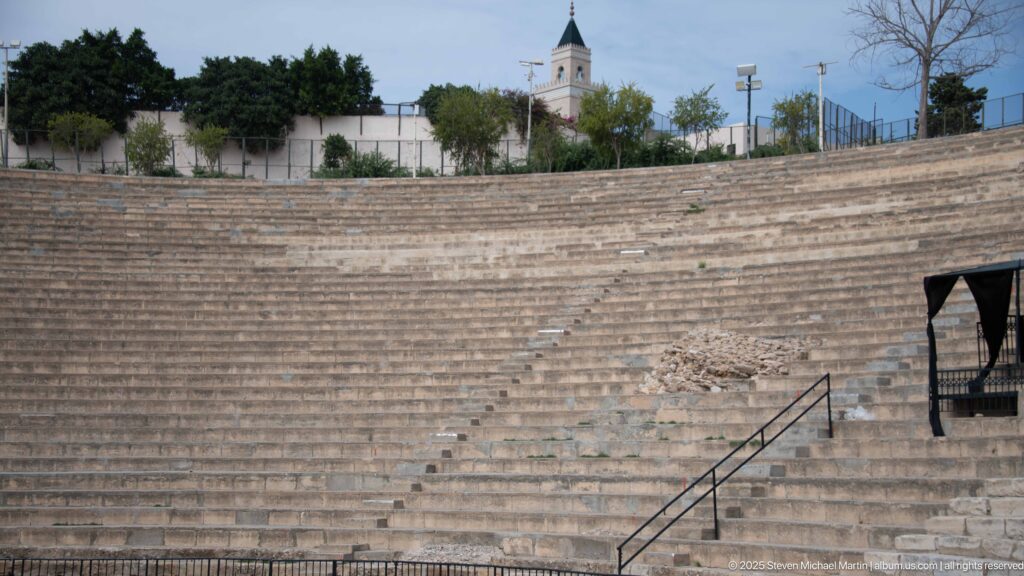Upper stone seating of the Roman Theatre of Carthage with repaired masonry, railings, trees, and distant mosque minaret.