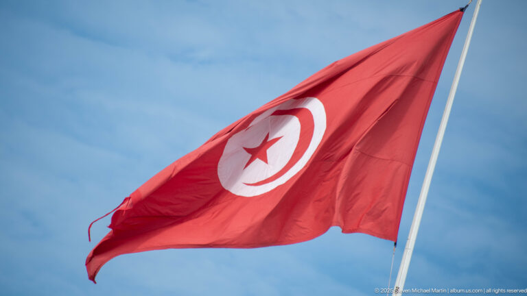 Red Tunisian flag with white disk, crescent, and star flying from a pole against blue sky.