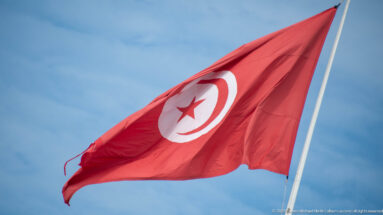 Red Tunisian flag with white disk, crescent, and star flying from a pole against blue sky.