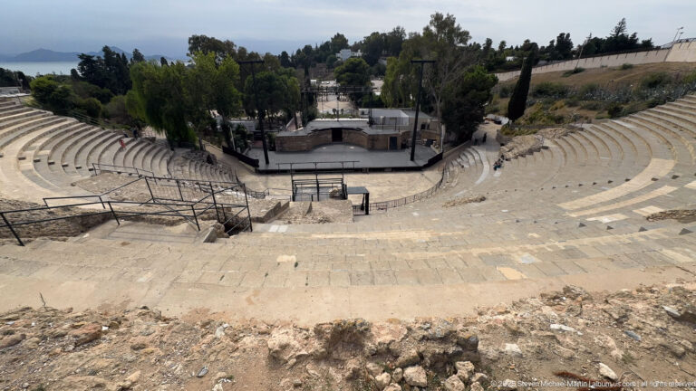 Centered high-angle view of the Roman Theatre of Carthage with curving stone seating around the stage.