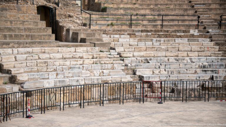 Close view of weathered and restored stone theatre seating with an access opening and black protective railings.