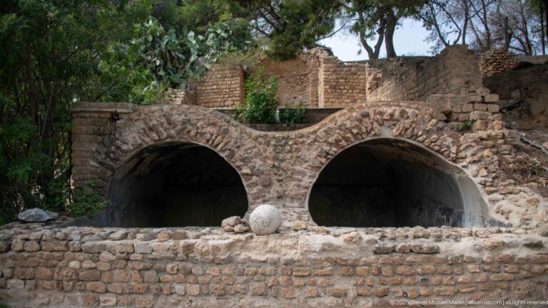 Two side-by-side barrel-vaulted openings in rough ancient masonry with ruins and trees behind.