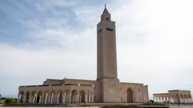 Square minaret of Malik ibn Anas Mosque above a long arcaded façade under a pale sky.