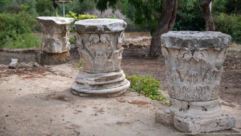 Three weathered stone capitals with acanthus carving standing beside the Roman archaeological remains at Carthage.