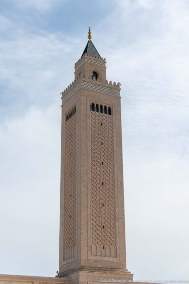 Upper portion of a square mosque minaret with patterned masonry, arched openings, and a green pyramidal roof.