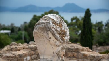 Weathered white marble head fragment with deeply carved hair, overlooking the Gulf of Tunis at Carthage.