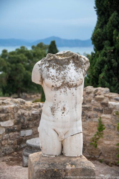 Headless and armless white marble male torso on a stone base with the Gulf of Tunis beyond.