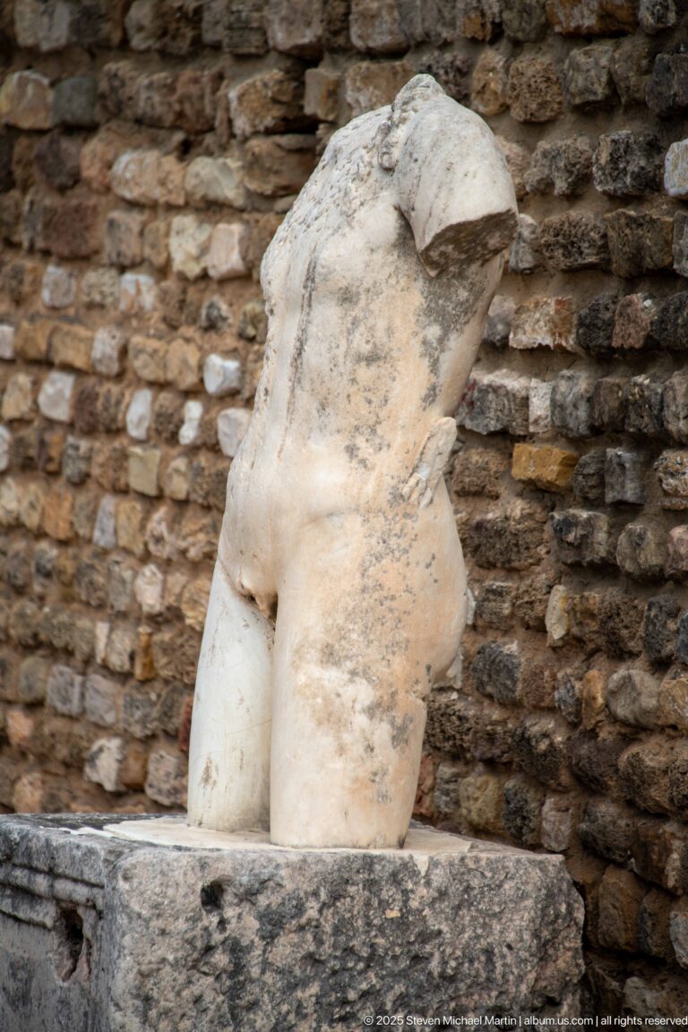 Headless white marble torso of a probable female figure displayed against rough ancient masonry.