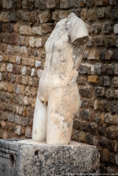 Headless white marble torso of a probable female figure displayed against rough ancient masonry.