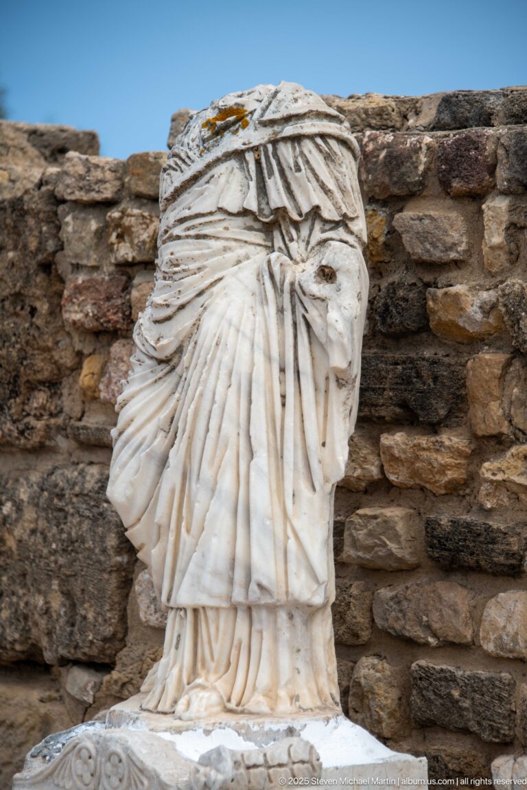 Fragmentary white marble statue with deeply carved layered drapery, missing its head and upper body.