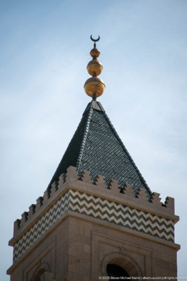 Green tiled pyramidal mosque roof topped by a stacked golden finial and crescent against the sky.