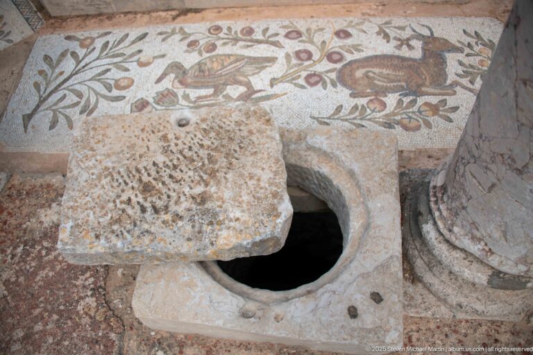 Circular stone-lined floor opening with a heavy partial cover beside a Roman bird mosaic and column base.