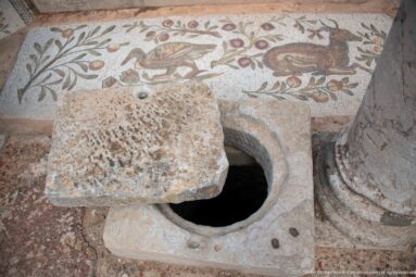 Circular stone-lined floor opening with a heavy partial cover beside a Roman bird mosaic and column base.