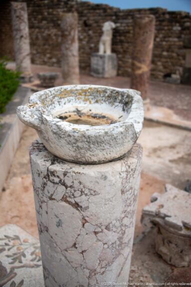 Shallow circular stone basin mounted on a short veined marble column within a Roman villa courtyard.