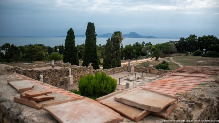 Elevated view across Roman villa ruins, columns, and reconstructed roofs toward the Gulf of Tunis and distant hills.
