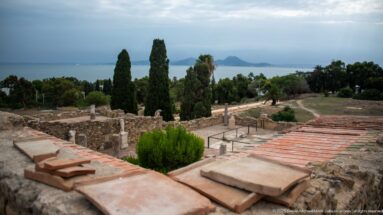 Elevated view across Roman villa ruins, columns, and reconstructed roofs toward the Gulf of Tunis and distant hills.