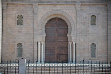 Horseshoe-arched wooden mosque entrance with pale carved stonework, slender columns, screened windows, and iron fence.