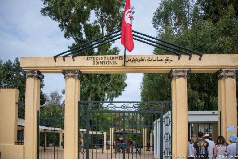 Bilingual entrance gateway for the Baths of Antoninus with French and Arabic site name beneath a Tunisian flag.