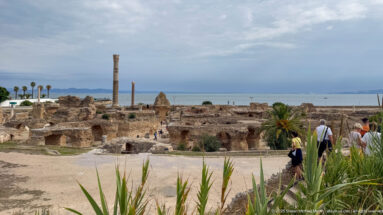 Wide elevated view of the Baths of Antoninus with vaulted ruins, standing columns, visitors, and the Mediterranean beyond.